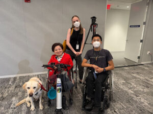 ID: (Left) to (Right) is Mia Ives-Rublee’s service dog, Mia Ives-Rublee, in a wheelchair, Casey Doherty, who is standing, and me (Huá Huī), who is in a wheelchair.